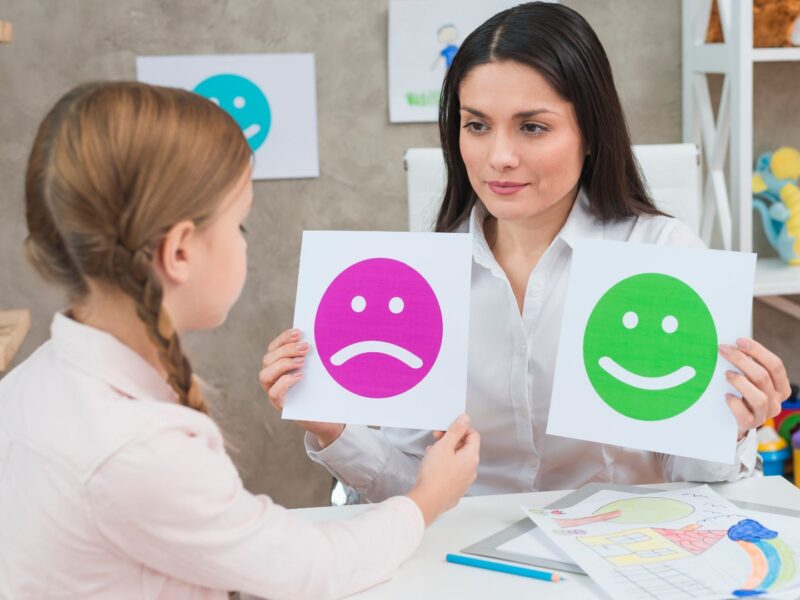 close-up-girl-choosing-sad-face-emoticons-paper-held-by-smiling-young-psychologist